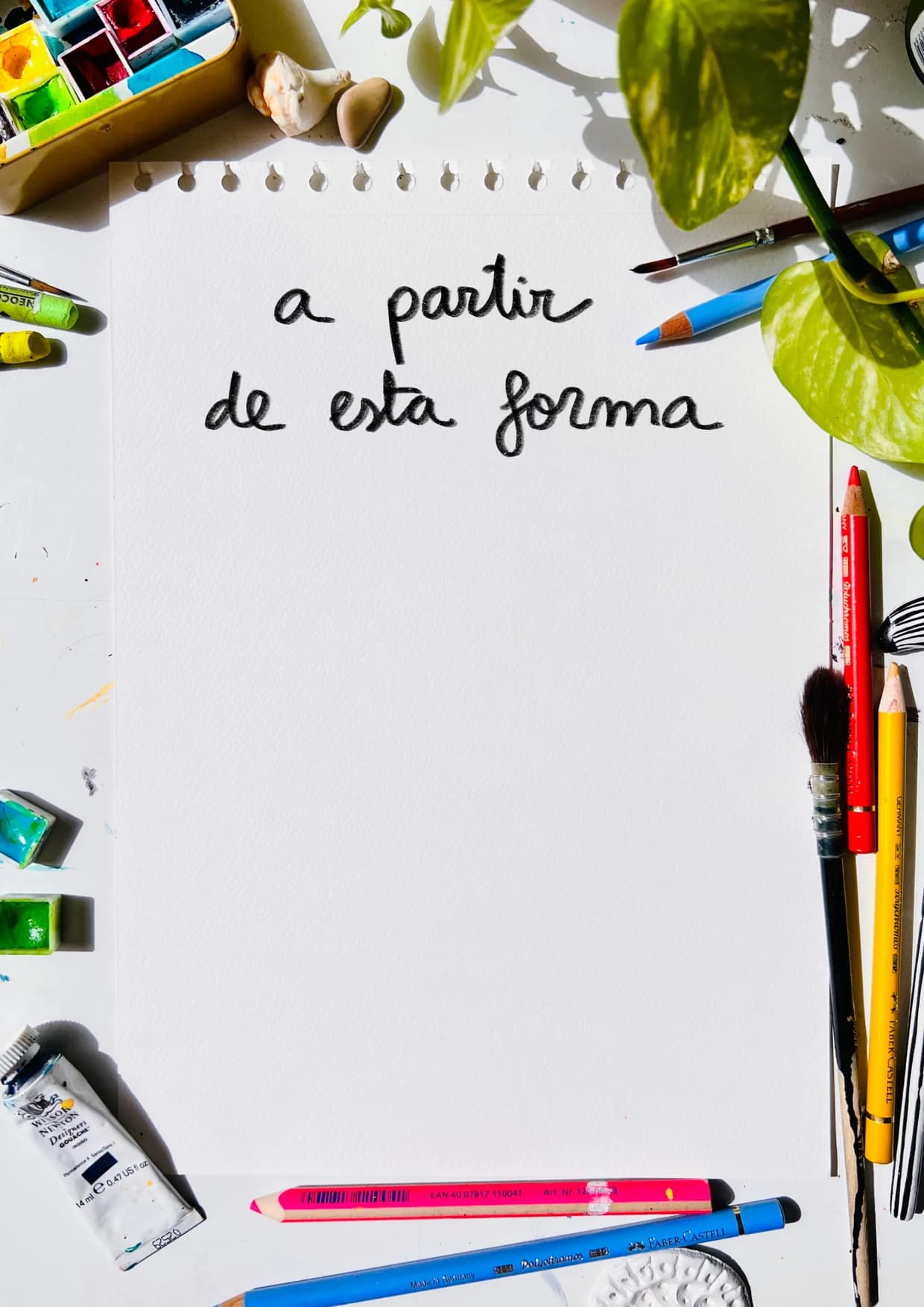 Hoja de papel rodeada de lápices de colores, pinceles, acuarelas y una planta. En la hoja, escrito a mano: «a partir de esta forma».